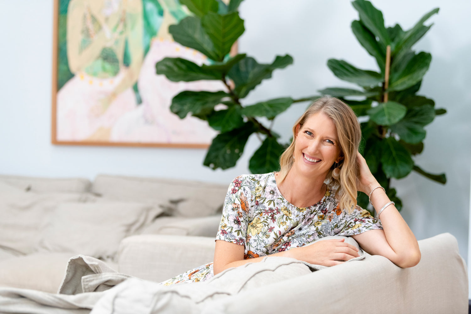 Anna, founder of Lighthouse Lane relaxing on a plush natural sofa wearing a floral dress. There is calming artwork and a fiddle leaf fig in the background.