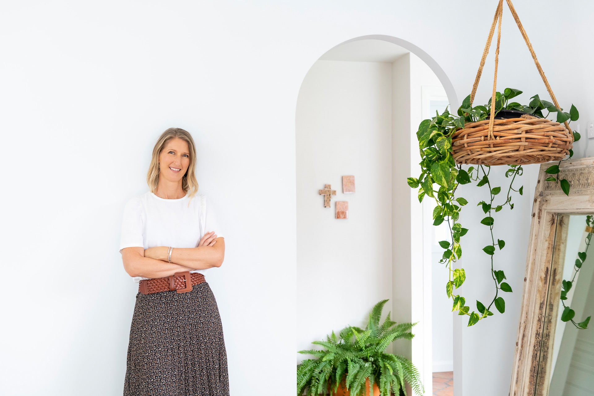 Anna the founder of Lighthouse Lane is standing with her arms casually crossed against a white wall. To her right is a hanging basket of green pothos and in the background is a lush fern.