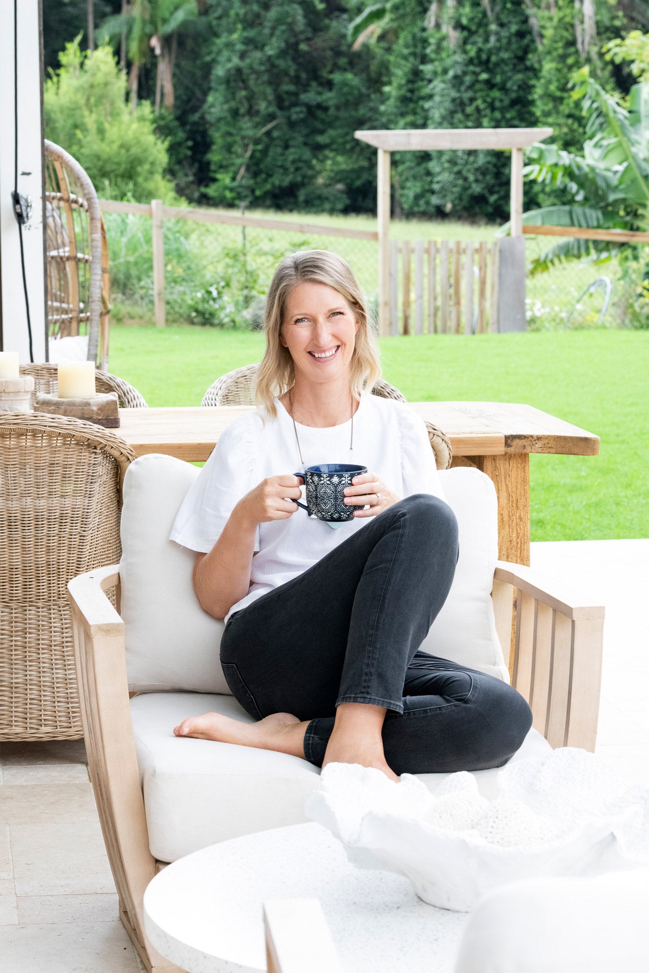 Lighthouse Lane founder Anna wears casual jeans and a tshirt. She is relaxed sitting in a timber outdoor chair holding a mug of tea. There is a lush garden in the background.