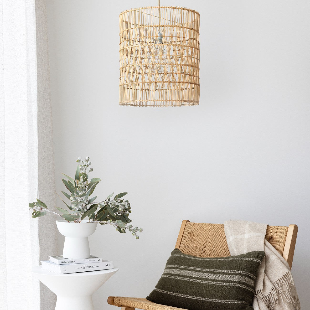 rattan pendant light shade. cylindrical light shade hanging above an occasional chair with an olive green cushion, and a vase of gum leaves.