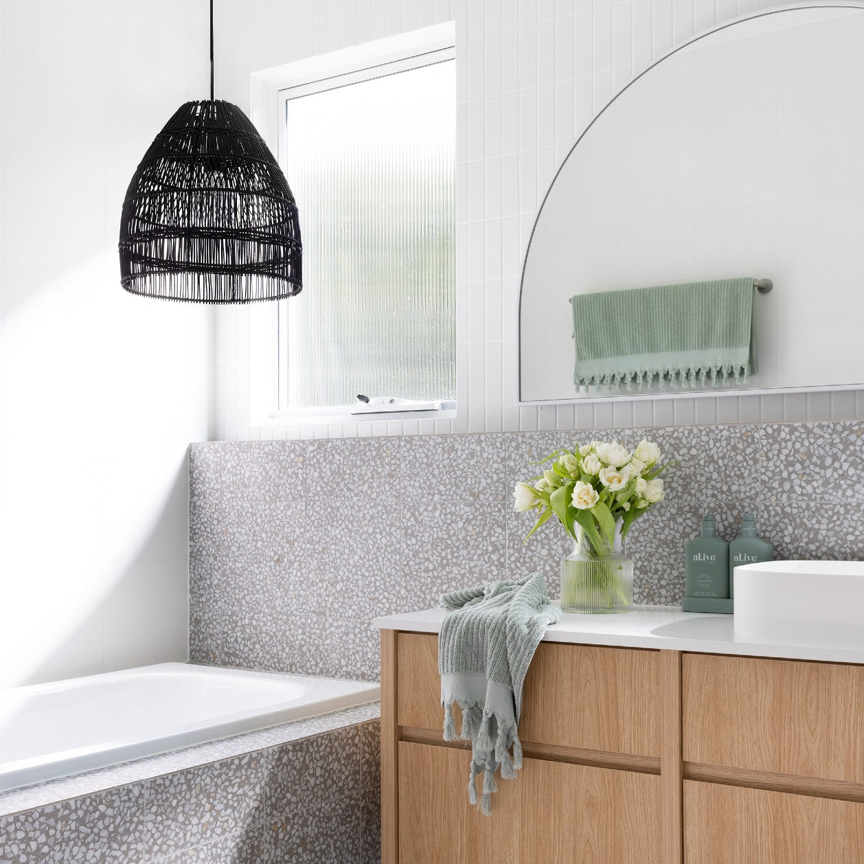 black rattan pendant light hangs above a bath in a grey terrazo tiled bathroom with accents of sage green.