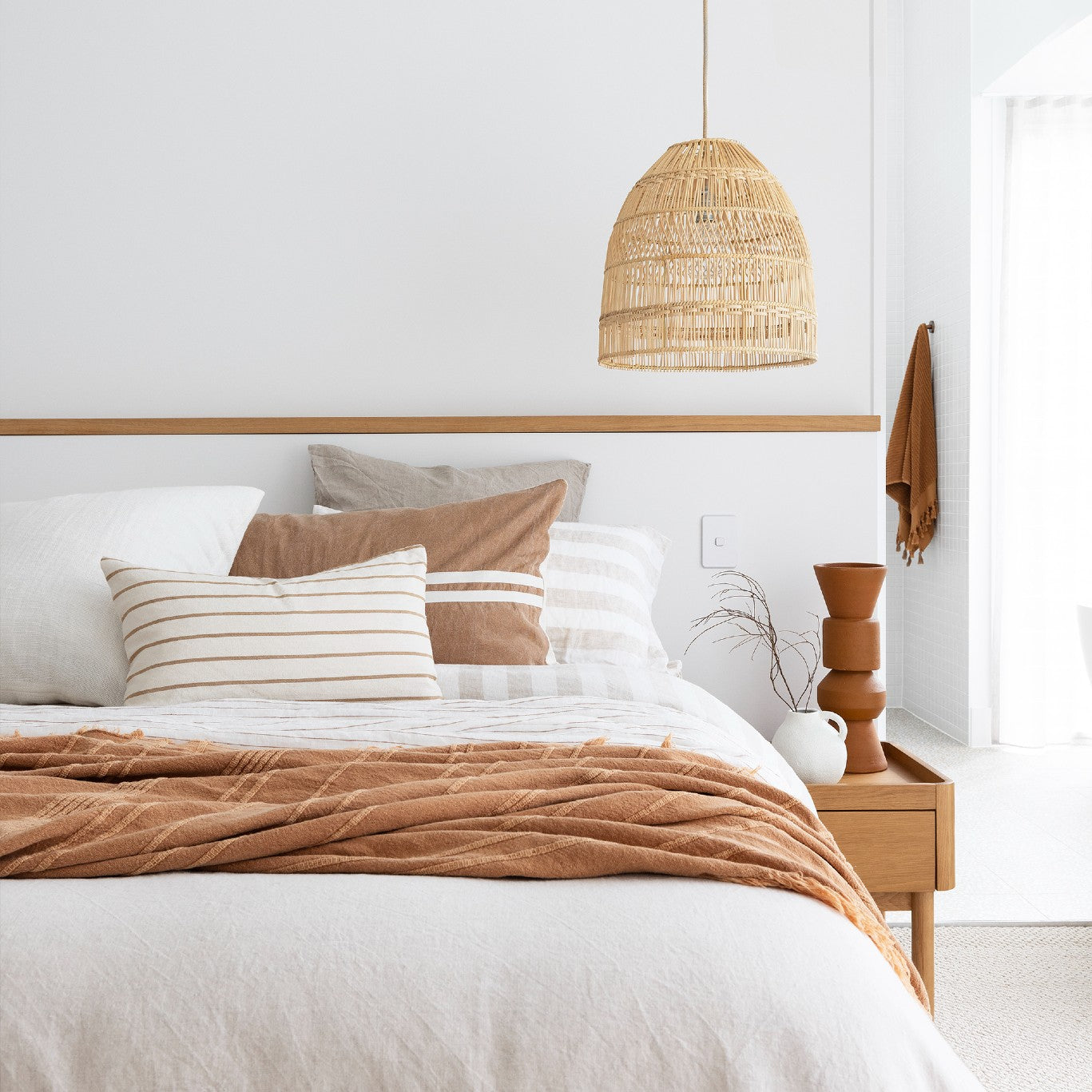 Coastal style pendant light hanging in a light filled bedroom. the bed is dressed in natural linen with a burnt orange throw and a matching towel hangs in the ensuite behind.