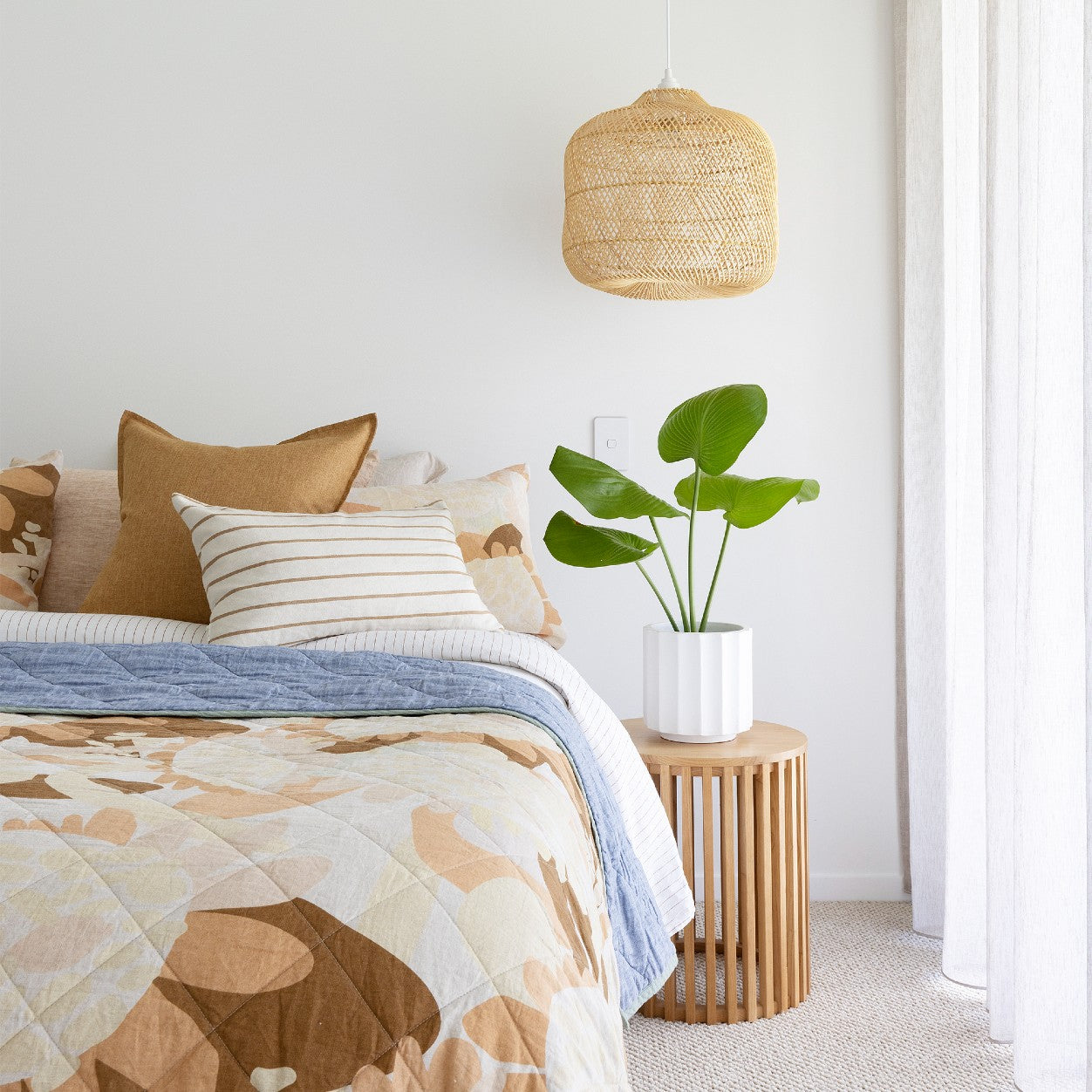 Rattan boho pendant light. Apple shaped rattan light used as a bedside pendant hanging next to retro inspired bed linen, a timber side table and a lush green plant.